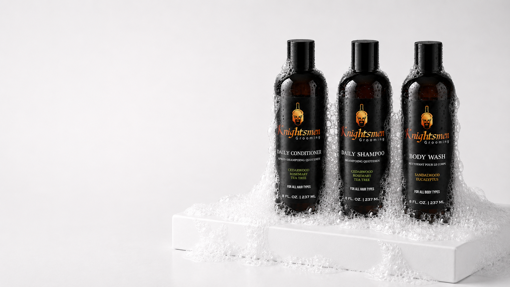 Three black bottles of Kingsmen Naturals grooming products (daily conditioner, daily shampoo, and body wash) standing in white foam on a white platform against a light gray background.