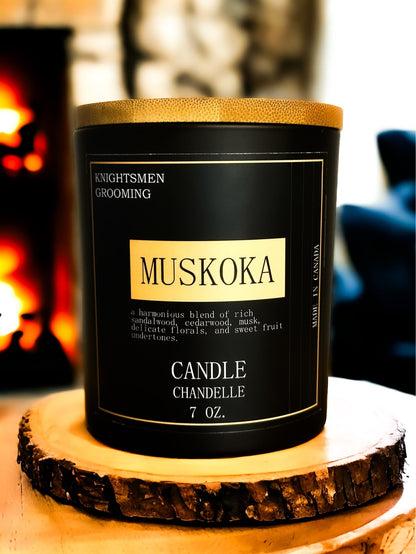 Muskoka candle in a black container with gold accents on a wooden coaster.